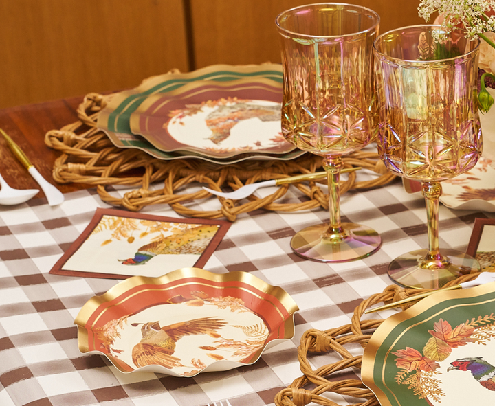 A table is set with Sophistiplate Woodland Harvest Appetizer/Dessert Bowls (8pk) featuring bird motifs, two iridescent glasses, and rattan placemats on a checkered tablecloth. A fall-themed napkin, disposable festive tableware, and white-handled cutlery are also visible.
