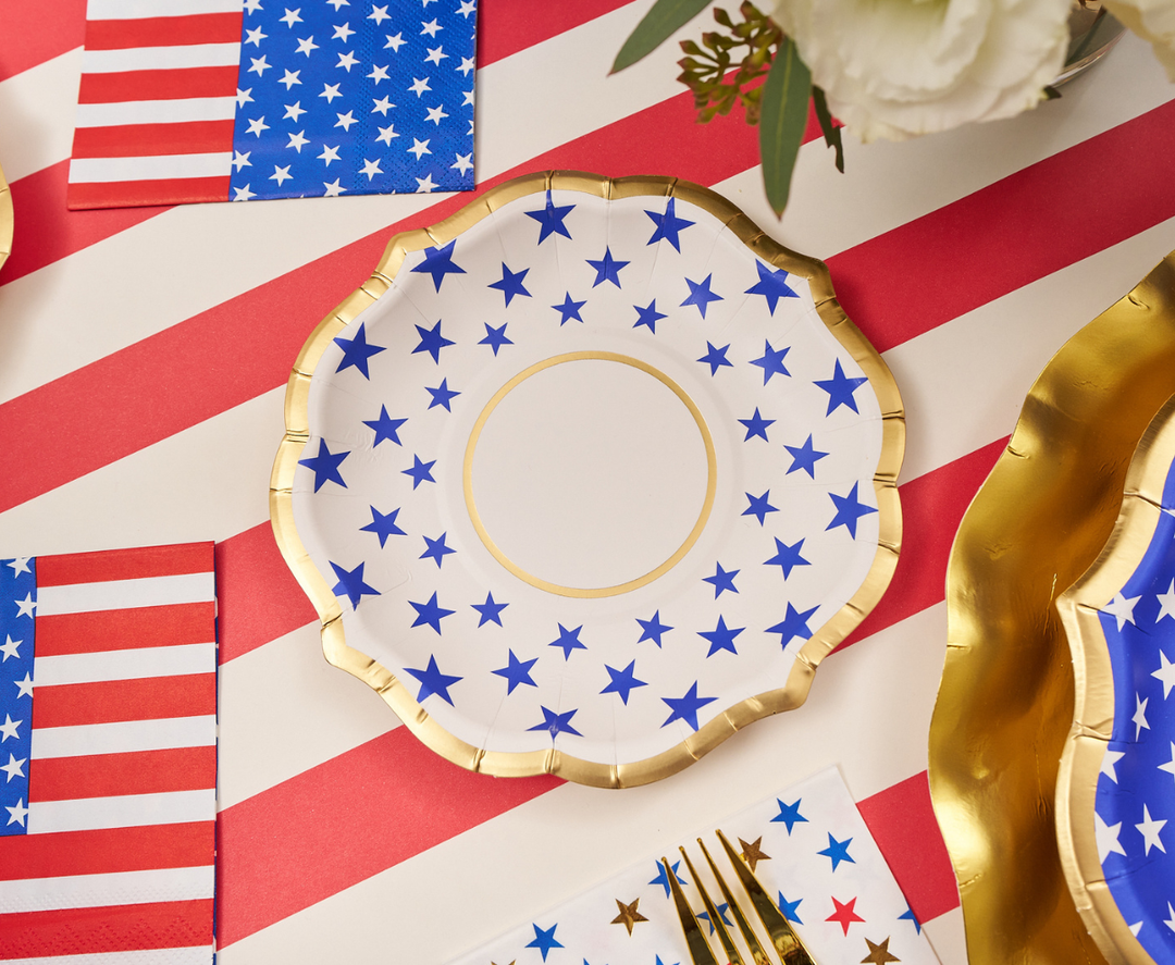 A decorative plate with blue stars and a gold rim sits on a red and white striped tablecloth, alongside Sophistiplate’s Party in the USA Appetizer/Dessert Bowls (8pk), American flag napkins, gold utensils, and a white flower arrangement.