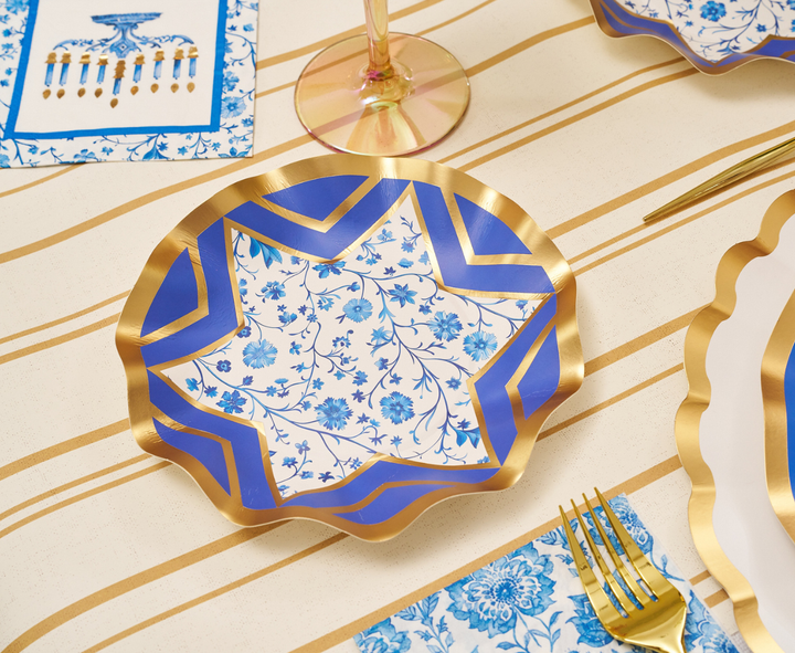 The Hanukkah Toile Table Setting by Sophistiplate features a decorative plate adorned with blue, gold, and floral patterns resting on a striped tablecloth. It is accompanied by a gold fork and knife, a floral napkin, and a wine glass. A partially visible Hanukkah-themed card adds to the scene, completing this festive tablescape with an elegant touch—perfect for the occasion with designer paper plates from Sophistiplate.