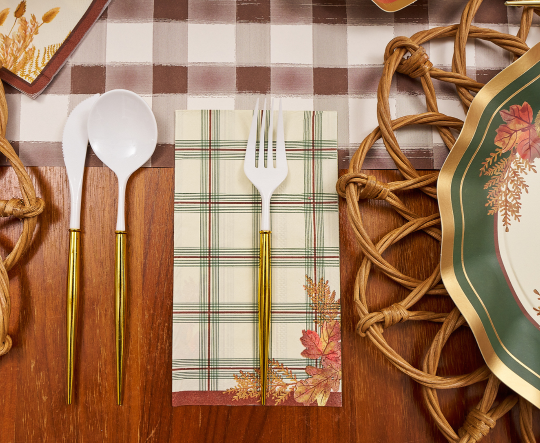 Sure! Here’s your rewritten sentence incorporating the product data: Sophistiplate Woodland Harvest Guest Towels are arranged elegantly beneath plastic gold-handled spoons, forks, and knives beside a fall-themed plate and rattan placemat, all set on a wooden table with a checkered table runner.
