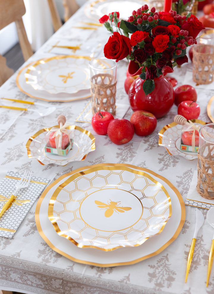 Elevate your Rosh Hashanah celebration with the Sophistiplate LLC “Rosh Tablescape for 16 Guests: White Honeybee,” featuring gold-accented, bee-themed designer disposable plates, yellow cutlery, apples, honey jar favors, woven glasses, and a red floral centerpiece on a patterned white tablecloth.