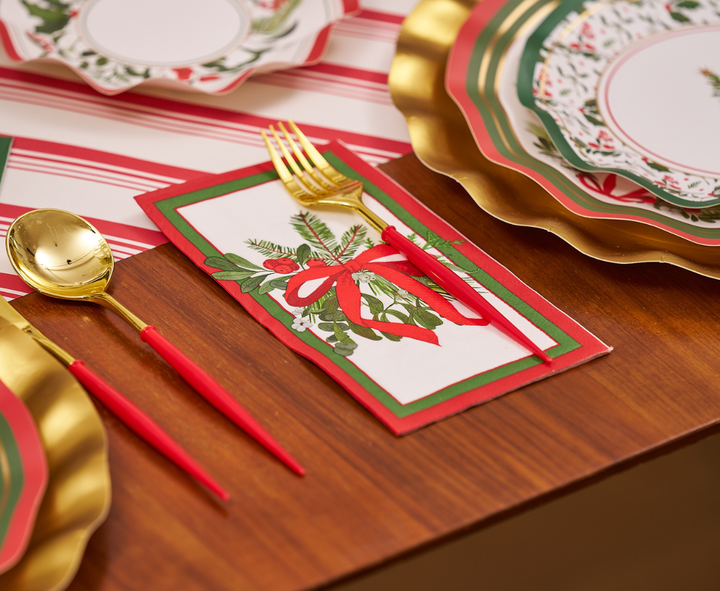Create a festive place setting featuring gold and red utensils, a napkin adorned with a holiday wreath and red bow, and decorative plates arranged on a wooden table with a red and white striped runner. Add an elegant touch to your tablescape by incorporating Sophistiplate Home for the Holidays Guest Towels (20pk) for that perfect holiday finishing detail.