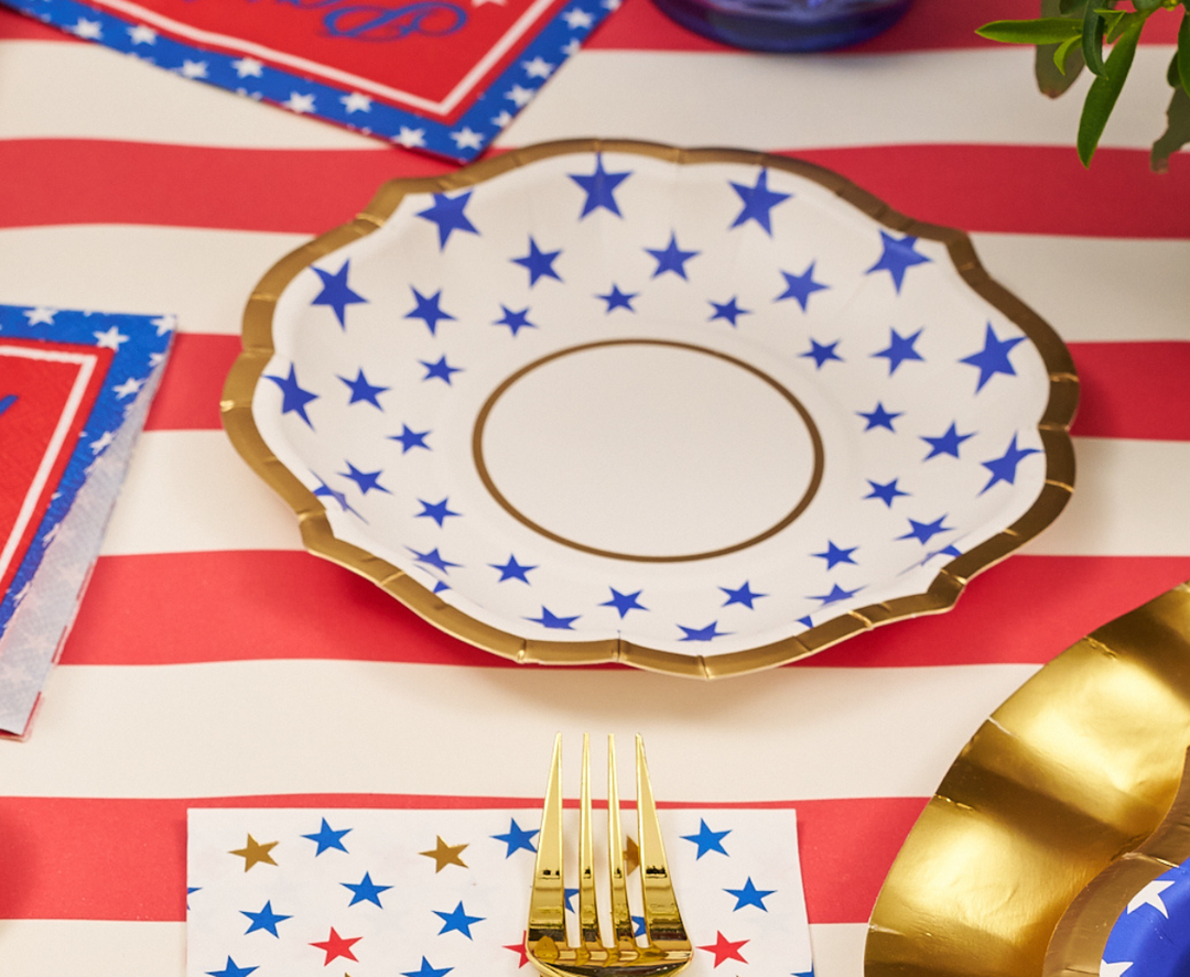 A decorative plate with blue stars and a gold rim sits on a red and white striped tablecloth, alongside Sophistiplate Party in the USA Appetizer/Dessert Bowls (8pk) and festive napkins with red, blue, and gold star patterns.