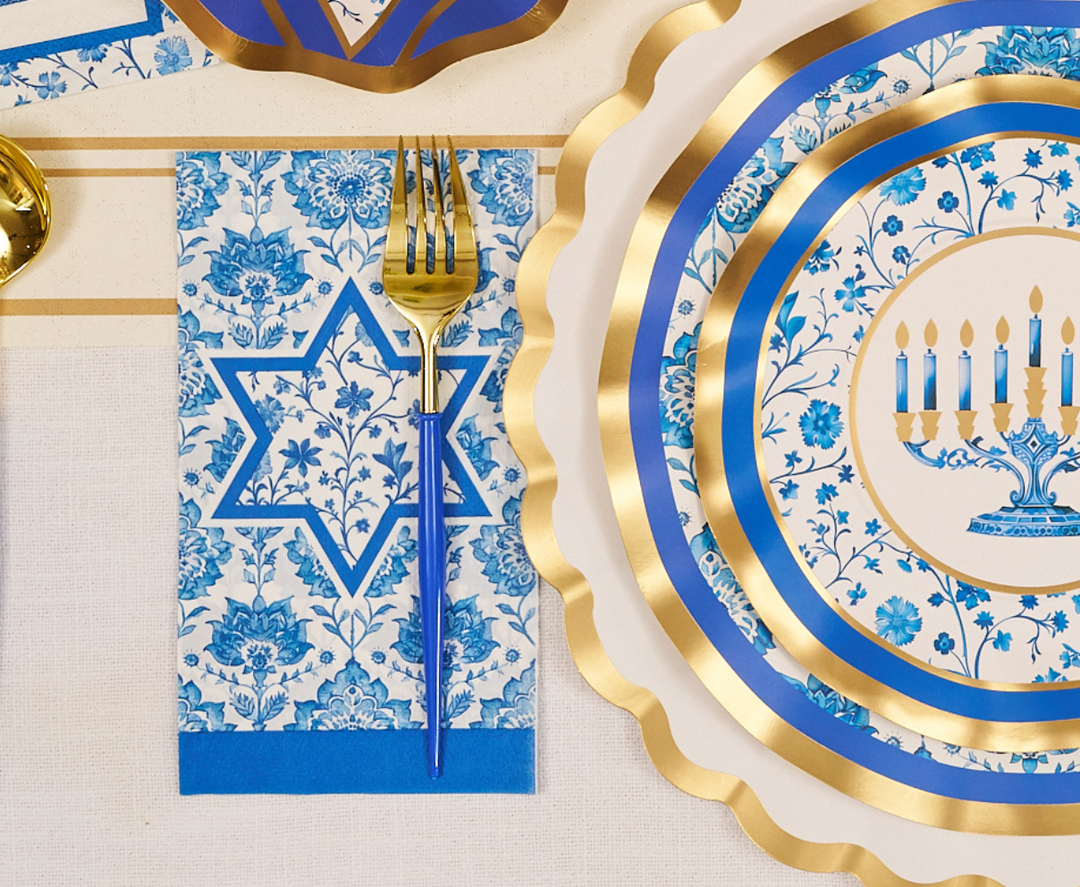 The Hanukkah Toile Table Setting by Sophistiplate creates a festive tablescape with blue and gold designer paper plates, coordinated napkins featuring a Star of David, elegant gold forks, and disposable plates decorated with menorahs and floral patterns.