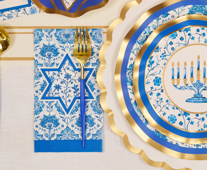 The Hanukkah Toile Table Setting by Sophistiplate creates a festive tablescape with blue and gold designer paper plates, coordinated napkins featuring a Star of David, elegant gold forks, and disposable plates decorated with menorahs and floral patterns.