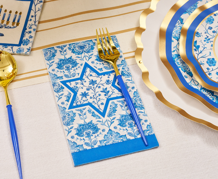 The Hanukkah Toile Table Setting by Sophistiplate showcases gold and blue utensils, a floral-patterned napkin adorned with a Star of David, a striped tablecloth, and elegant blue and gold-edged designer paper plates—ideal for celebrating Hanukkah in style.