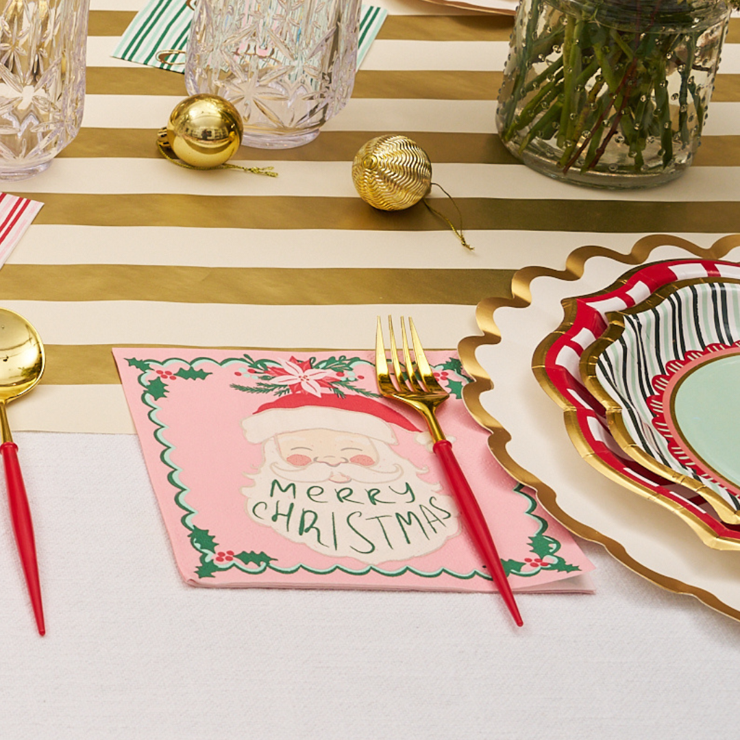 Celebrate the season with a festive table featuring a gold-striped runner, colorful plates and paper tableware, gleaming gold cutlery, sparkling Christmas ornaments, and Sugarplum’s Tis the Season Lunch Napkins (20pk)—pink napkins decorated with Santa and ‘Merry Christmas’ text. Glasses and a vase of flowers complete this joyful holiday gathering setup.
