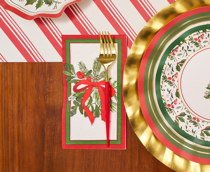 A gold fork rests on a Sophistiplate Home for the Holidays Guest Towel napkin, adorned with a red ribbon and greenery, placed next to a holiday-themed plate featuring red, green, and gold patterns on a wooden table.