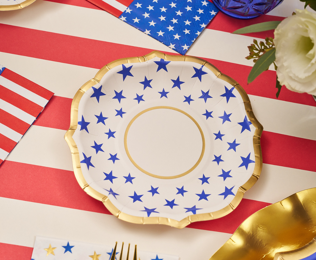 A decorative plate with blue stars and a gold rim sits on a red and white striped tablecloth, surrounded by Sophistiplate Party in the USA Appetizer/Dessert Bowls (8pk), American flag napkins, a gold fork, and a white flower arrangement.