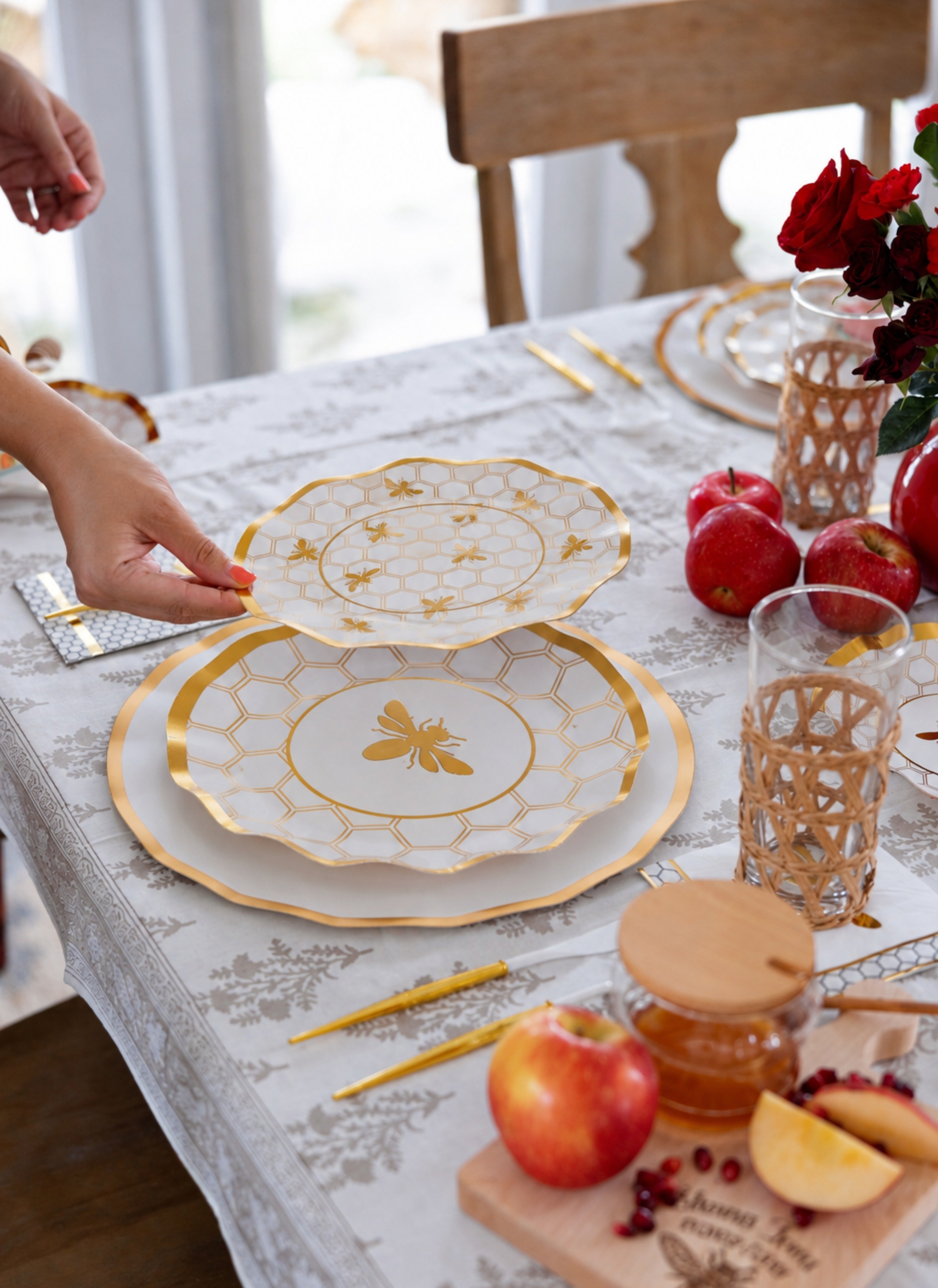 A hand lifts a plate from the Sophistiplate White Honeybee Table Setting, featuring bee-themed gold rim disposable tableware, apples, honey, cutlery, glasses, and a vase of red flowers arranged on a white patterned tablecloth.