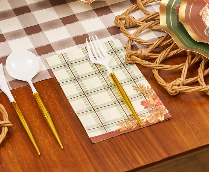 A white plastic spoon and fork with gold handles rest on a Sophistiplate Woodland Harvest Guest Towel, featuring a fall-themed plaid design, placed next to woven placemats and stacked green-rimmed plates on a wooden table.