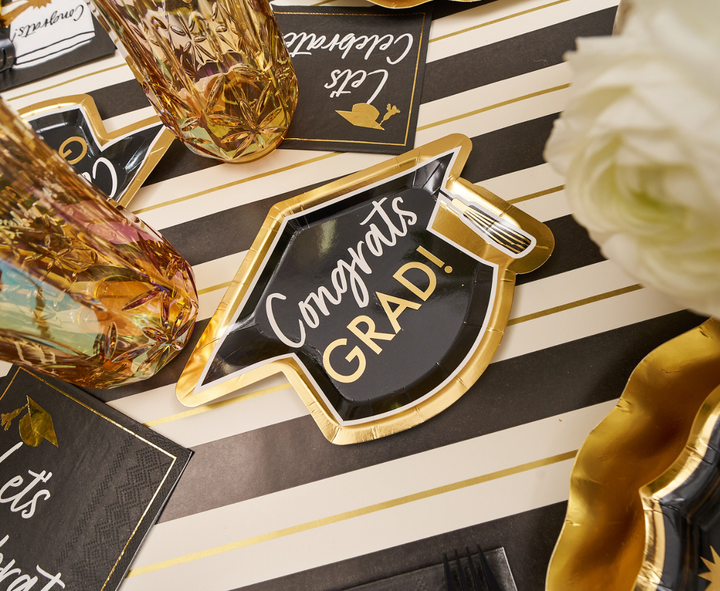 The Sophistiplate Classic Grad Table Setting features a black and gold graduation cap-shaped plate with "Congrats GRAD!", gold-rimmed glasses, napkins, and white flowers on a striped tablecloth.