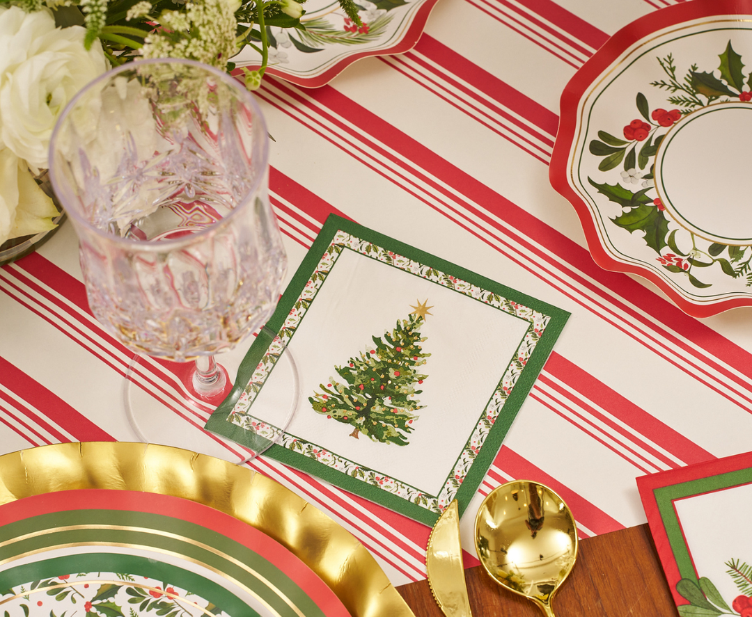 Create an elegant and festive atmosphere at your next gathering with the Home for the Holidays Table Setting by Sophistiplate, featuring a crystal glass, gold spoon, designer disposable plates, and a napkin adorned with a Christmas tree—all arranged on a red and white striped tablecloth for the perfect holiday tablescape.