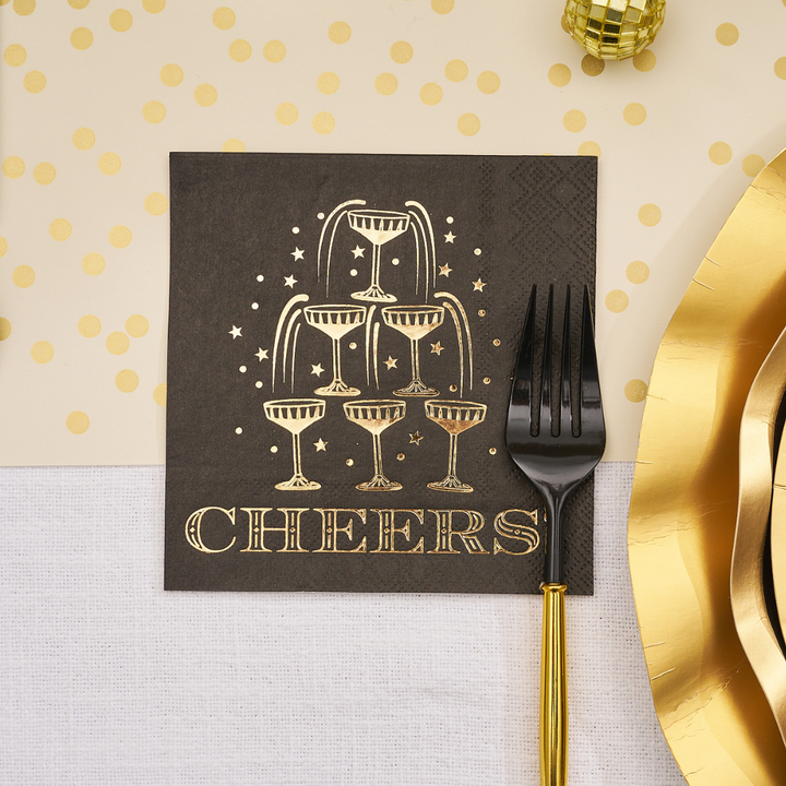Midnight Sparkle Paper Cocktail Napkin Cheers/26pk, a black square napkin featuring a gold foil champagne tower and the word CHEERS, set beside a black and gold fork for a New Year’s Eve party.