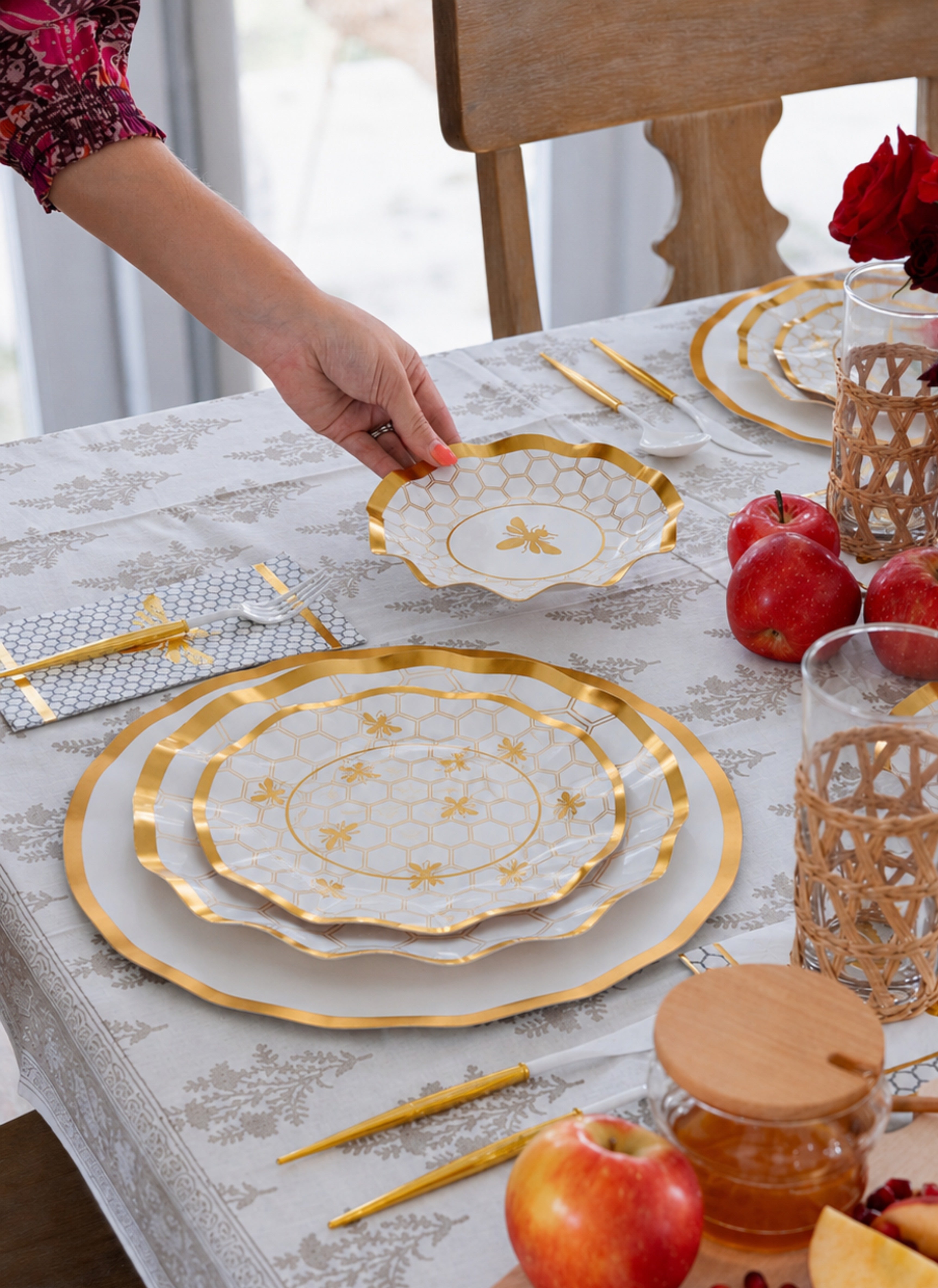 A hand sets the Sophistiplate LLC “Rosh Tablescape for 16 Guests: White Honeybee” gold-trimmed bowl, featuring a bee motif, on a table accented with matching designer disposable plates, red apples, woven glasses, and gold utensils atop a patterned tablecloth—creating an elegant Rosh Hashanah setting.