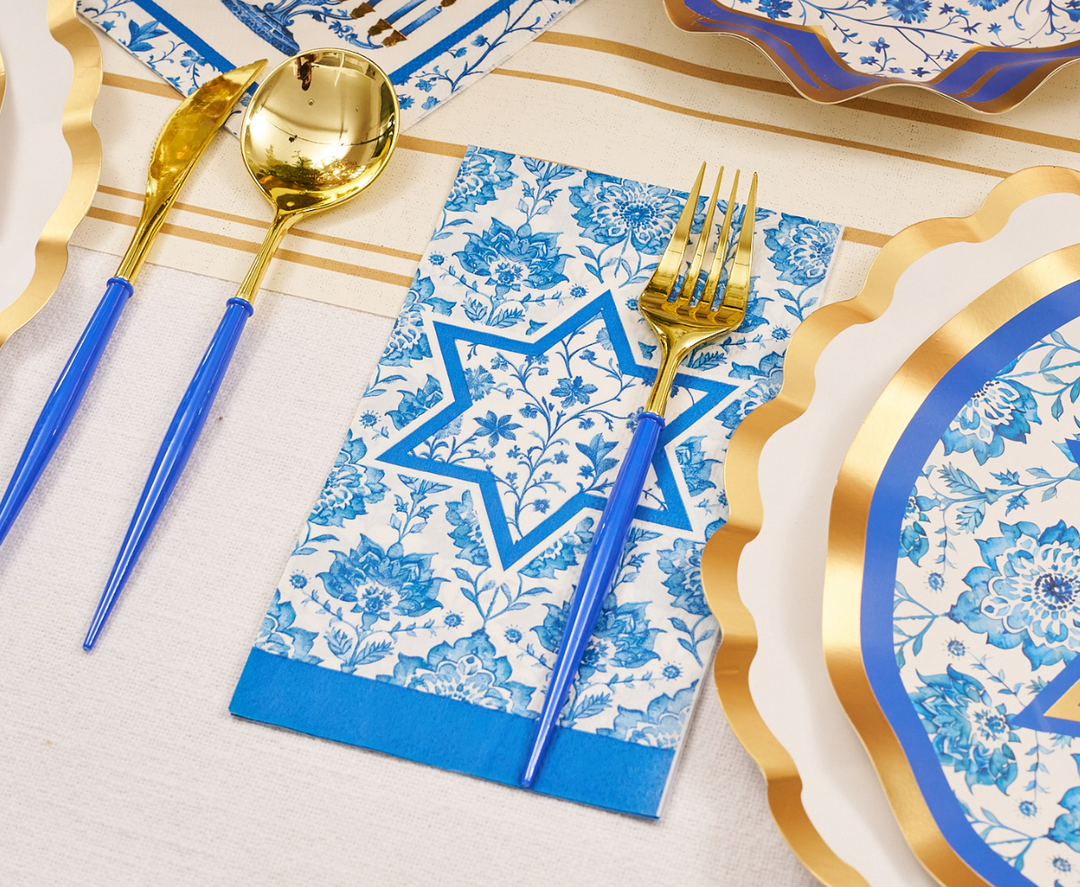The Hanukkah Toile Table Setting by Sophistiplate features blue and gold utensils resting on a white tablecloth, a floral napkin adorned with a blue Star of David, and designer paper plates with scalloped gold edges—arranged together for a festive Hanukkah tablescape.