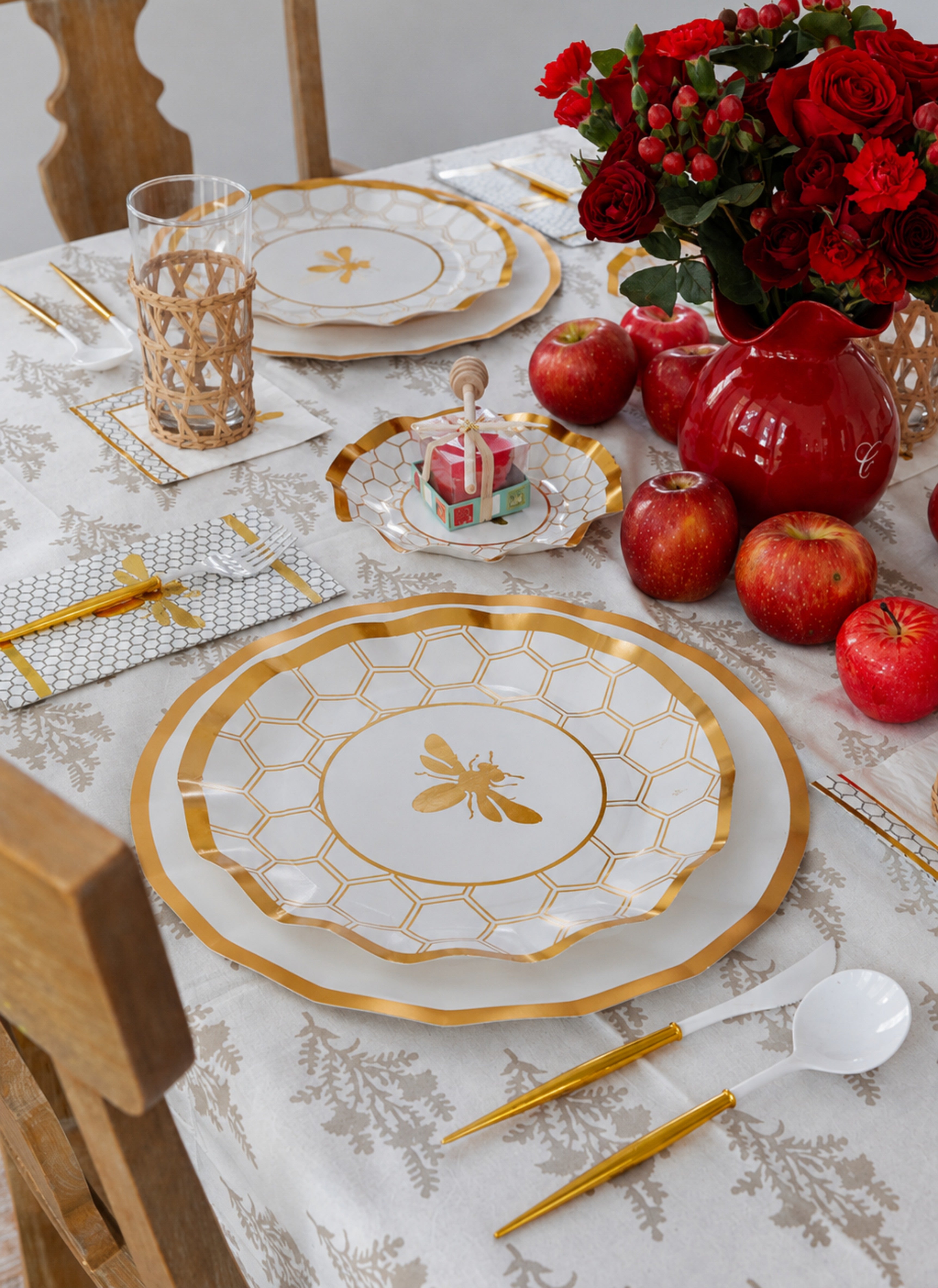 A festive table set for a meal features Sophistiplate LLC’s Rosh Tablescape for 16 Guests: White Honeybee, showcasing elegant gold and white bee-themed heavy duty paper plates. The setting is complemented by apples, gold utensils, a glass, and a vase filled with red roses—perfect for creating a celebratory Rosh Hashanah ambiance. A small birthday cake with candles is displayed on one of the stylish disposable party plates to complete the scene.