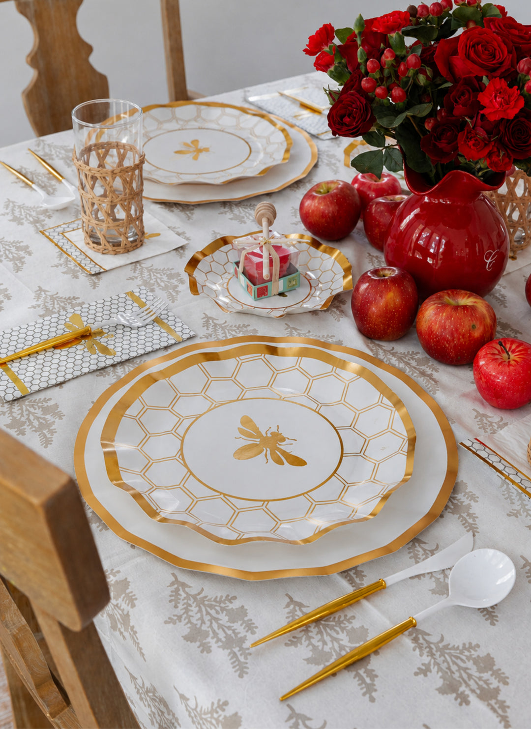 A festive table set for a meal features Sophistiplate LLC’s Rosh Tablescape for 16 Guests: White Honeybee, showcasing elegant gold and white bee-themed heavy duty paper plates. The setting is complemented by apples, gold utensils, a glass, and a vase filled with red roses—perfect for creating a celebratory Rosh Hashanah ambiance. A small birthday cake with candles is displayed on one of the stylish disposable party plates to complete the scene.