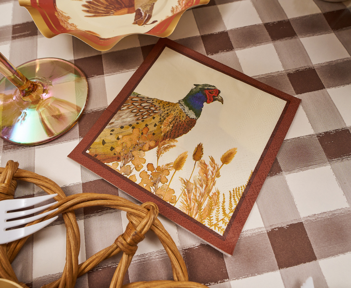 A Woodland Harvest Table Setting by Sophistiplate features a decorative napkin with an illustrated pheasant, arranged atop a brown-and-white checkered tablecloth and accompanied by a glass with an iridescent base, festive autumn party tableware, and a woven placemat set with white cutlery.