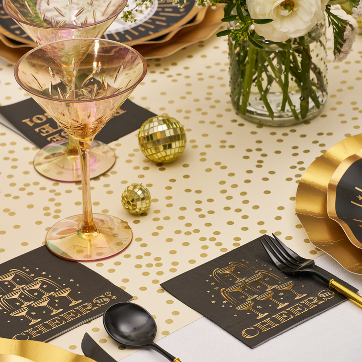 Sophistiplate Midnight Sparkle Paper Cocktail Napkin Cheers/26pk, featuring black napkins with a gold illustration of stacked champagne glasses and the word CHEERS, set on a gold-dotted tablecloth for a New Year’s Eve party.
