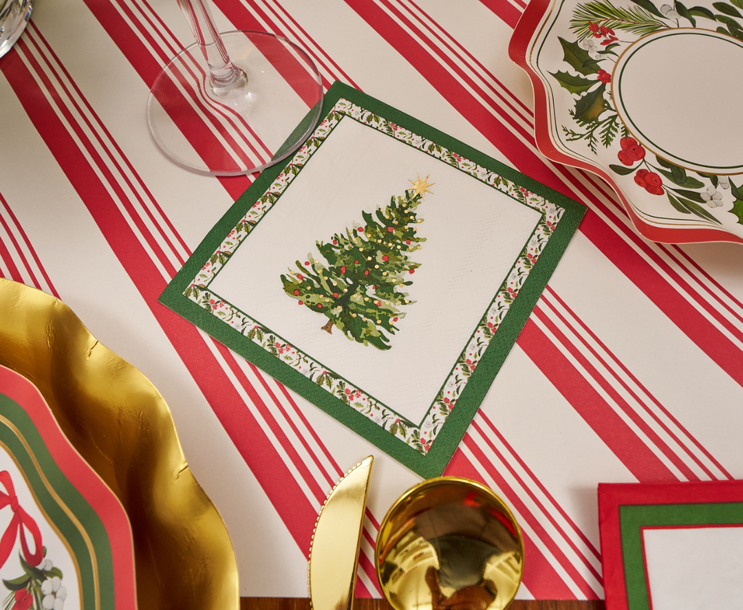 Create a festive holiday tablescape with the Home for the Holidays Table Setting by Sophistiplate, featuring a red and white striped tablecloth, gold utensils, napkins adorned with decorated Christmas trees, and designer disposable plates showcasing holly and greenery—perfect for elevating your Christmas party tableware.