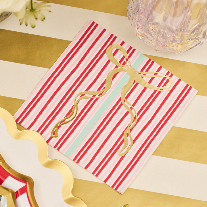 The Sophistiplate Tis the Season Cocktail Napkins (26pk) feature festive red, pink, and white stripes with a gold ribbon and bow design. Perfect for Christmas parties or holiday gatherings, these decorative napkins add a cheerful touch when placed on a gold-and-white striped surface next to a clear glass and scalloped plate.