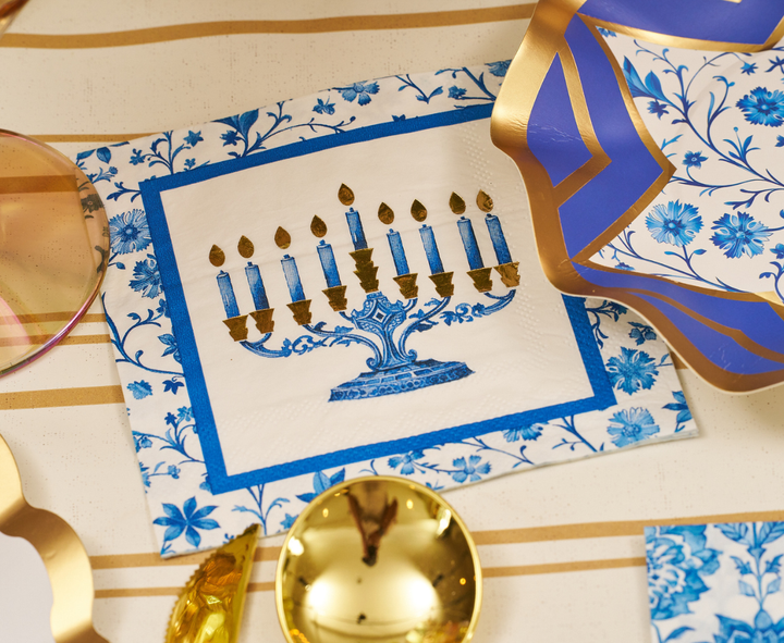 The Hanukkah Toile Table Setting by Sophistiplate features a decorative napkin with a blue and gold menorah design, complemented by festive elements such as blue floral plates, gold utensils, and a striped tablecloth.