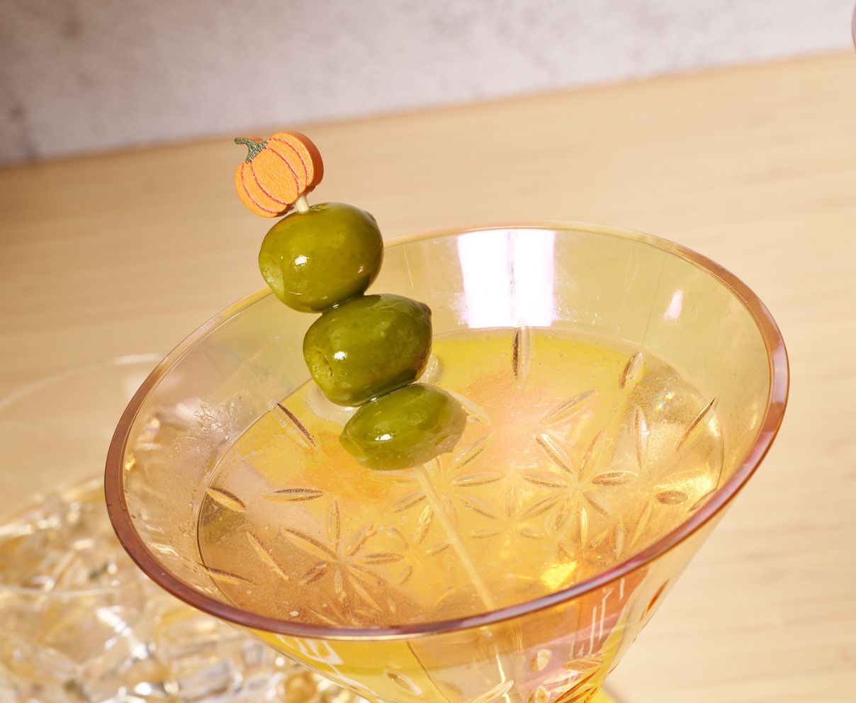 A close-up of a cocktail in a patterned glass, garnished with three green olives on Simply Serv'd Pumpkin Large Cocktail Picks/30pk. The drink rests on a light wooden surface.