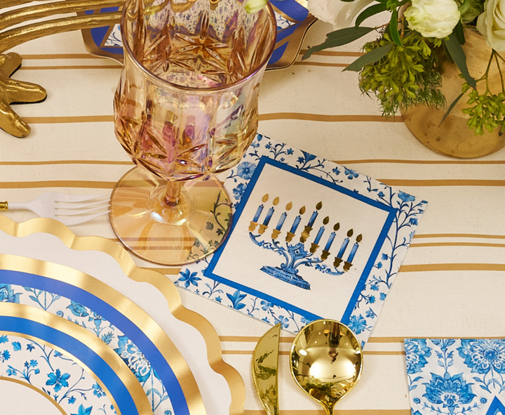 The Hanukkah Toile Table Setting by Sophistiplate features elegant gold utensils, a pink decorative glass, designer paper plates with blue and gold trim, and a napkin showcasing a blue menorah with six lit candles and one shamash. The festive tablescape is beautifully arranged and accented by white flowers presented in a gold vase.