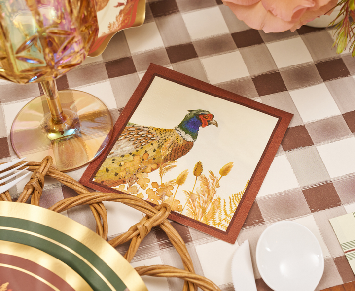 The Woodland Harvest Table Setting by Sophistiplate features a decorative napkin adorned with a vibrant pheasant motif, elegantly placed atop a checkered tablecloth and accented by a gold-rimmed plate, woven placemat, glassware, and seasonal floral arrangements—creating an inviting scene that beautifully complements designer disposable plates and autumn party tableware.