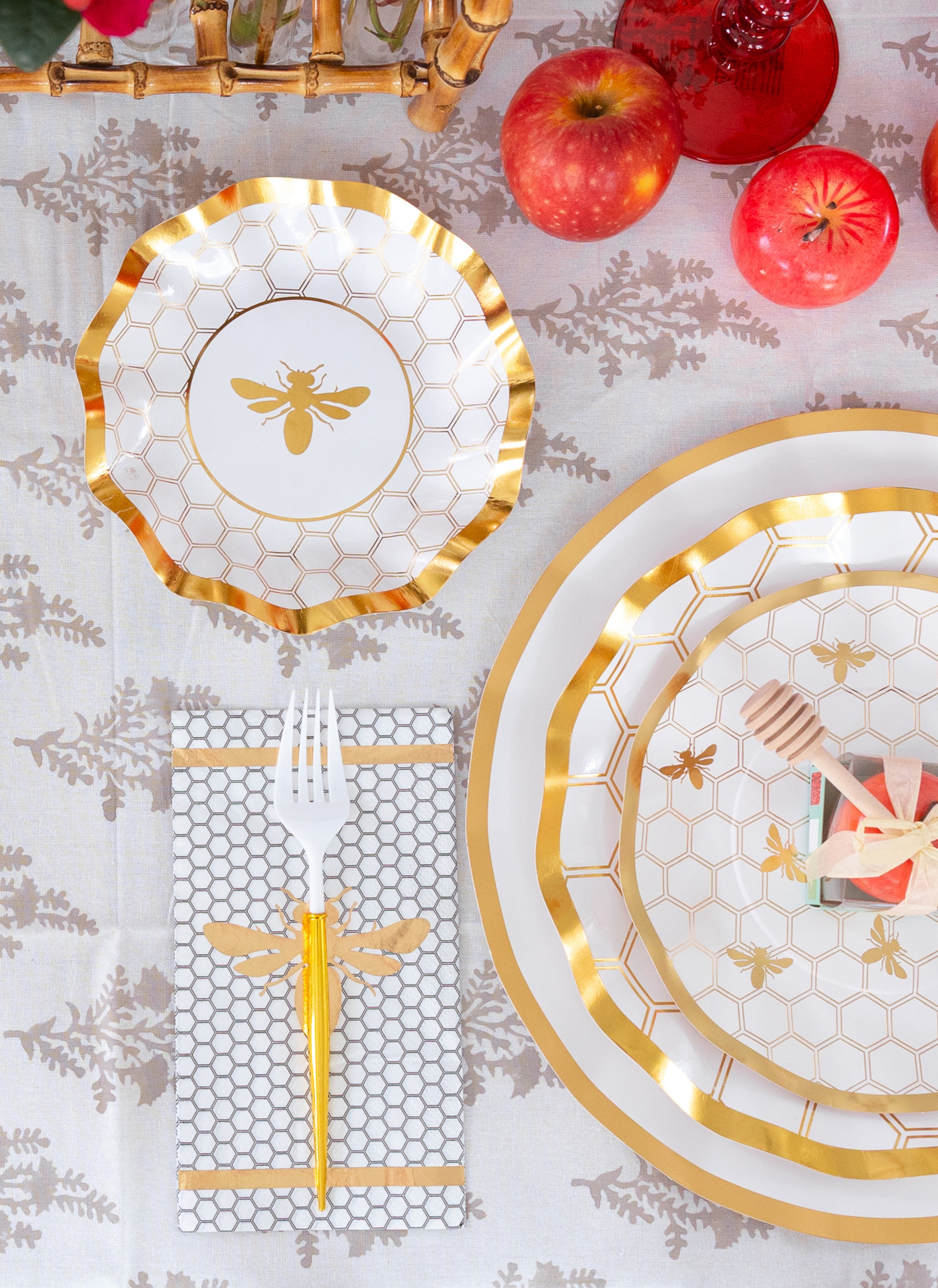 Create a stunning Rosh Hashanah table with the Sophistiplate LLC “Rosh Tablescape for 16 Guests: White Honeybee” set, featuring gold-rimmed heavy duty paper plates with honeycomb patterns, an elegant gold bee napkin, gold forks, coordinating pink-wrapped utensils, fresh apples, and red drinks—all beautifully arranged on a white and gray floral tablecloth.