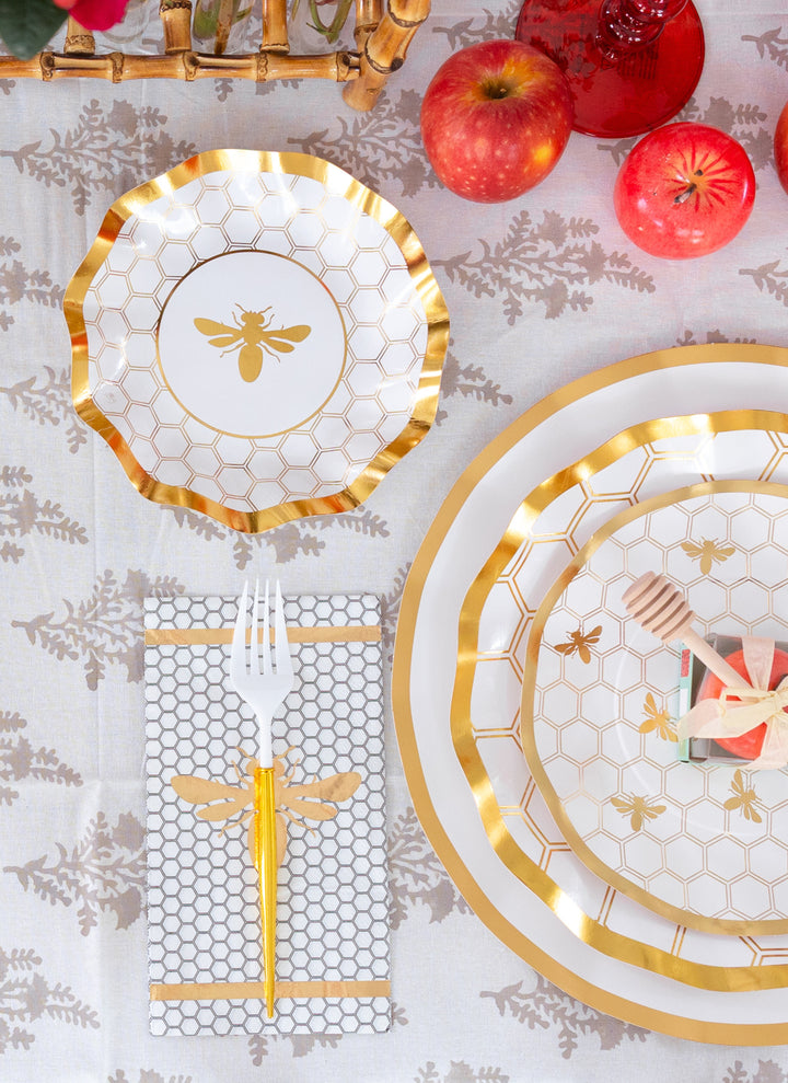 Create a stunning Rosh Hashanah table with the Sophistiplate LLC “Rosh Tablescape for 16 Guests: White Honeybee” set, featuring gold-rimmed heavy duty paper plates with honeycomb patterns, an elegant gold bee napkin, gold forks, coordinating pink-wrapped utensils, fresh apples, and red drinks—all beautifully arranged on a white and gray floral tablecloth.