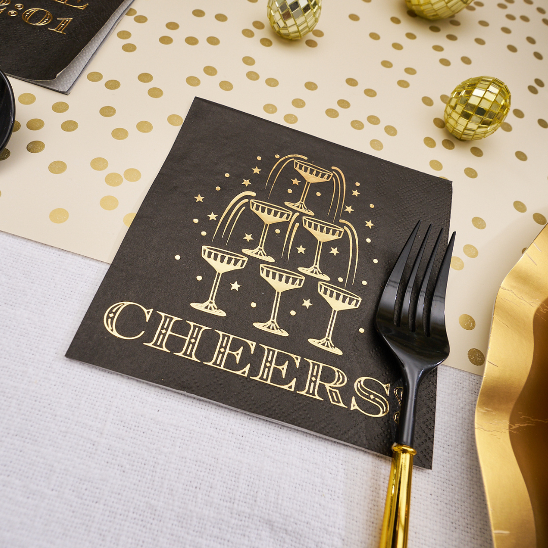 Midnight Sparkle Paper Cocktail Napkin Cheers/26pk with a gold foil illustration of a champagne tower and the word CHEERS printed below, set on a table next to a black fork, perfect for a New Year’s Eve party.