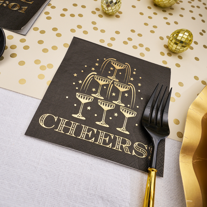 Midnight Sparkle Paper Cocktail Napkin Cheers/26pk with a gold foil illustration of a champagne tower and the word CHEERS printed below, set on a table next to a black fork, perfect for a New Year’s Eve party.