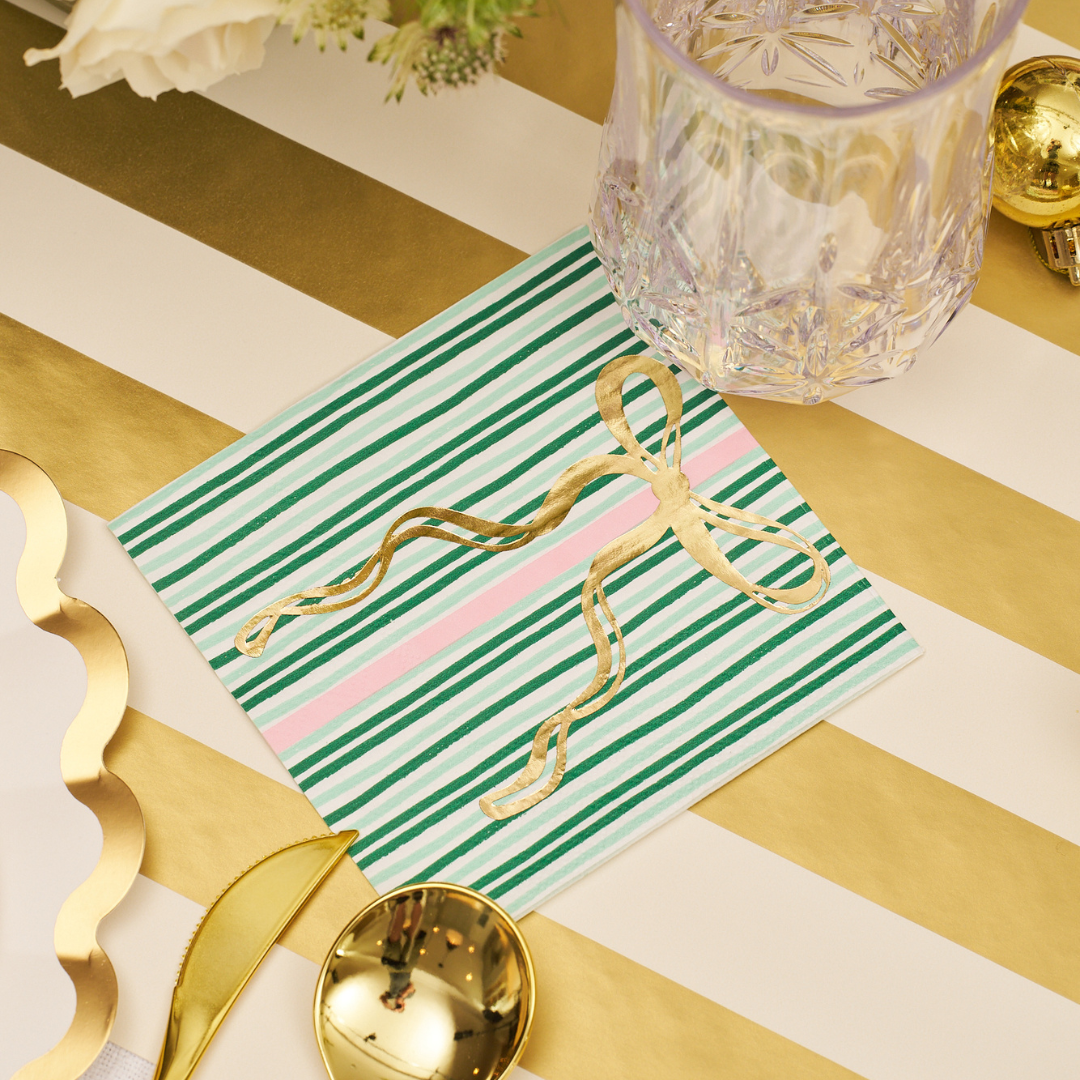 A Tis the Season Cocktail Napkin by Sophistiplate rests on a gold and white striped table beside a crystal glass, gold spoon, and decorative plate, accented with a gold ribbon bow.