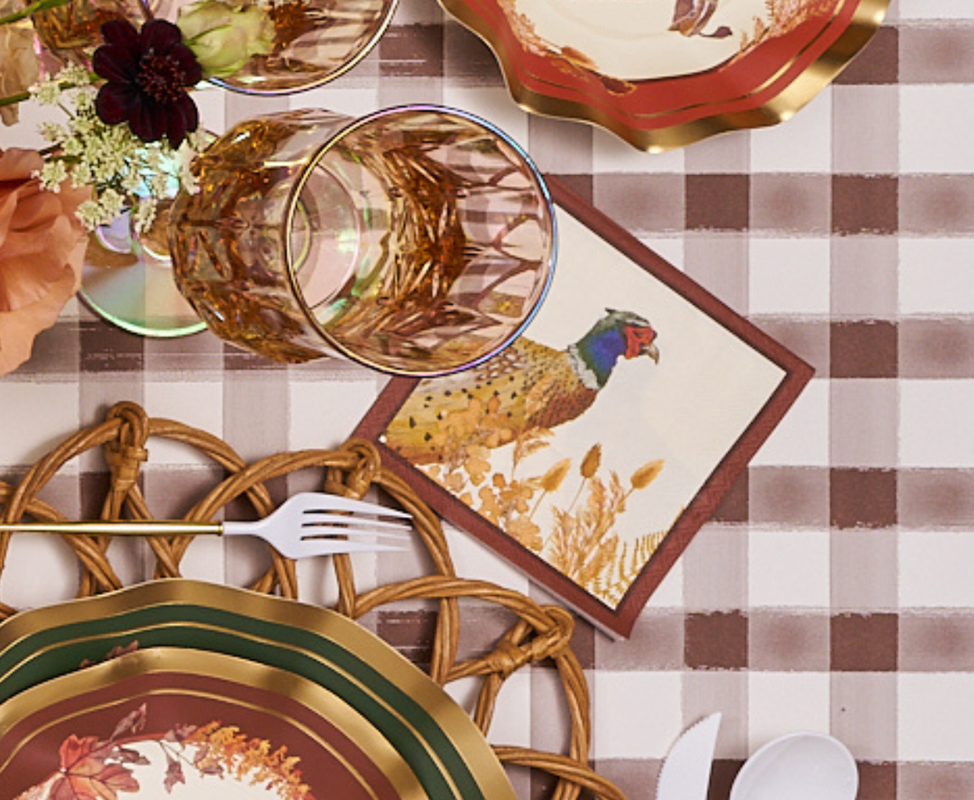 The Sophistiplate Woodland Harvest Table Setting, featuring plates, gold-rimmed glasses, a fork, and a pheasant-themed napkin arranged on a checkered tablecloth with floral accents, creates the perfect autumn party tableware display.