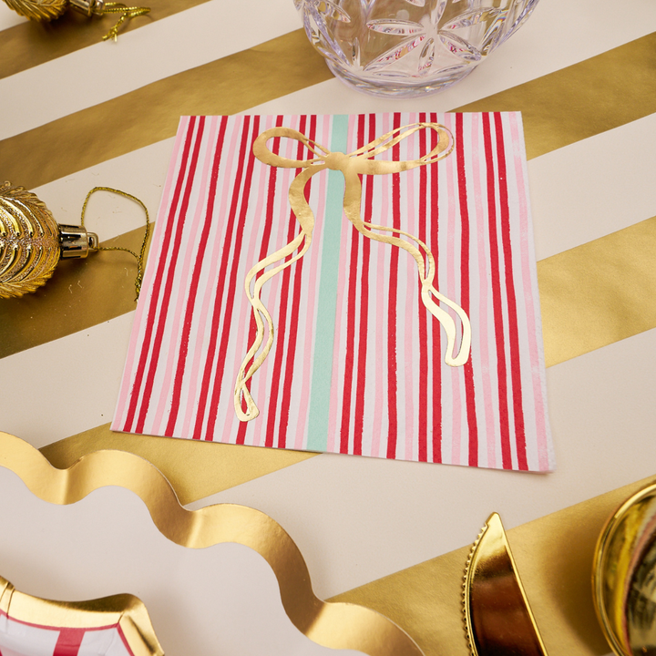 A “Tis the Season” Cocktail Napkin by Sophistiplate, featuring red, white, and light blue stripes with a shiny gold bow design, is displayed on a gold-striped table beside gold ornaments and a crystal glass.