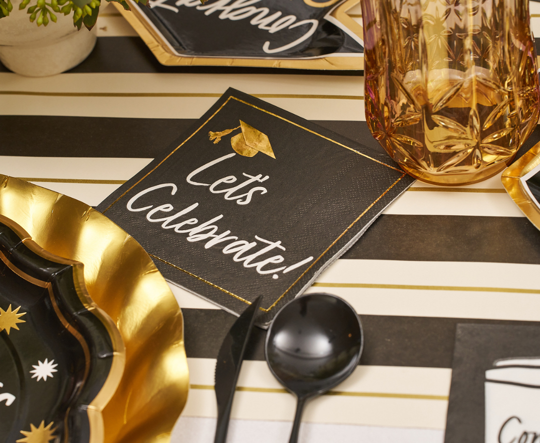 The Classic Grad Table Setting by Sophistiplate includes black "Let's Celebrate!" napkins with a gold cap, gold-edged plates, black utensils, and a pink glass on a striped tablecloth—perfect for stylish graduation parties.