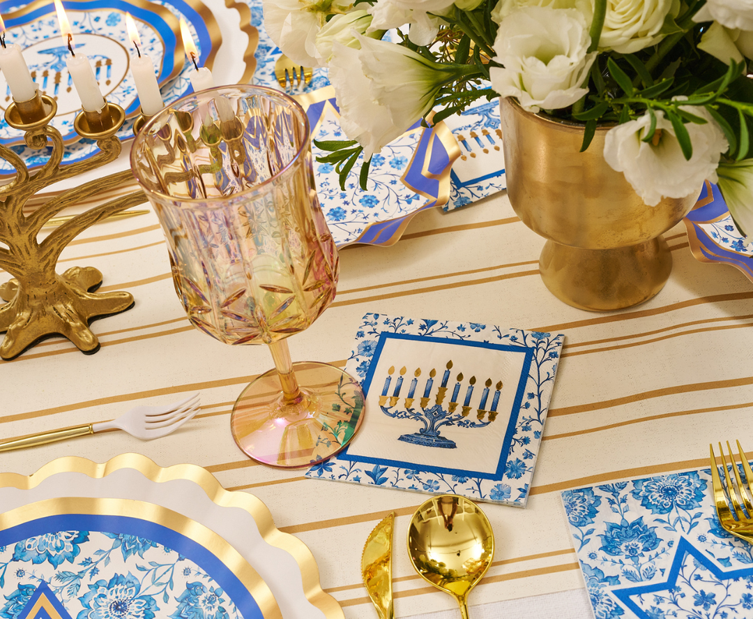Celebrate Hanukkah in style with the Sophistiplate Hanukkah Toile Table Setting, featuring blue and gold designer paper plates, gold utensils, a pink glass, a menorah napkin, and a gold vase with white flowers. A menorah centerpiece completes the festive atmosphere.