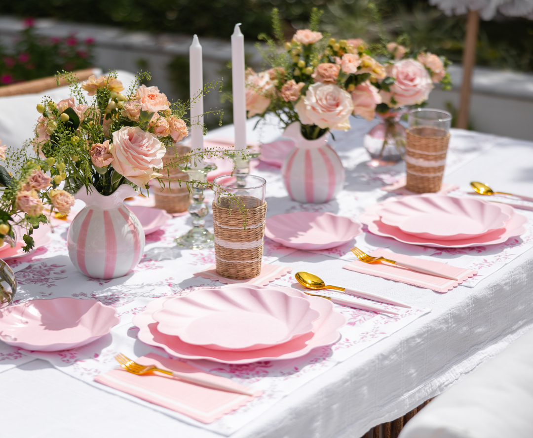 The Eco-Friendly Table Setting- Blush by Sophistiplate LLC features pink scalloped plates, gold cutlery, woven glasses, floral placemats, vases of blush roses, and white candles for an elegant, feminine outdoor table.