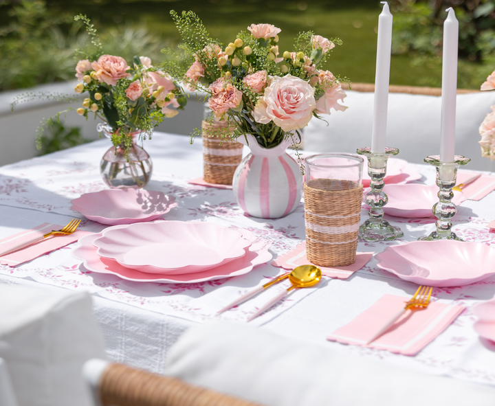 The Eco-Friendly Table Setting- Blush by Sophistiplate LLC features blush scalloped plates, gold cutlery, pink napkins, floral vases, white candles, and woven tumblers arranged on a white and pink tablecloth for a chic outdoor meal.