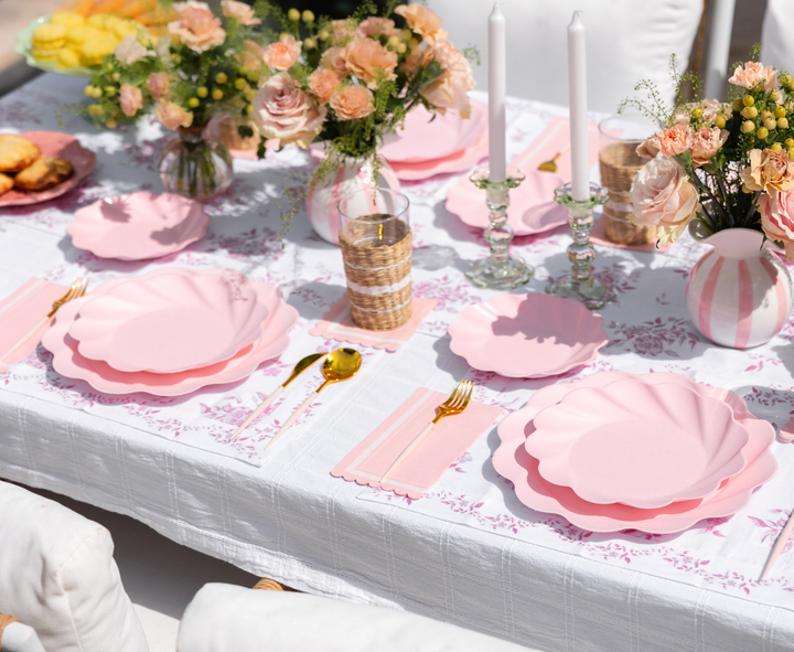 The Eco-Friendly Table Setting- Blush by Sophistiplate LLC features disposable plates, gold utensils, pink napkins, floral centerpieces, and white candles on a white tablecloth with a delicate pink pattern—perfect for elegant outdoor dining.