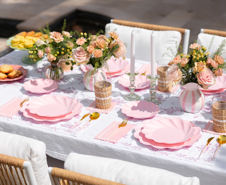 A spring table features Sophistiplate LLC’s Eco-Friendly Table Setting- Blush, gold flatware, wicker glasses, floral napkins, candles, vases with pink and white blooms, plus plates of cookies and pastries on the side.