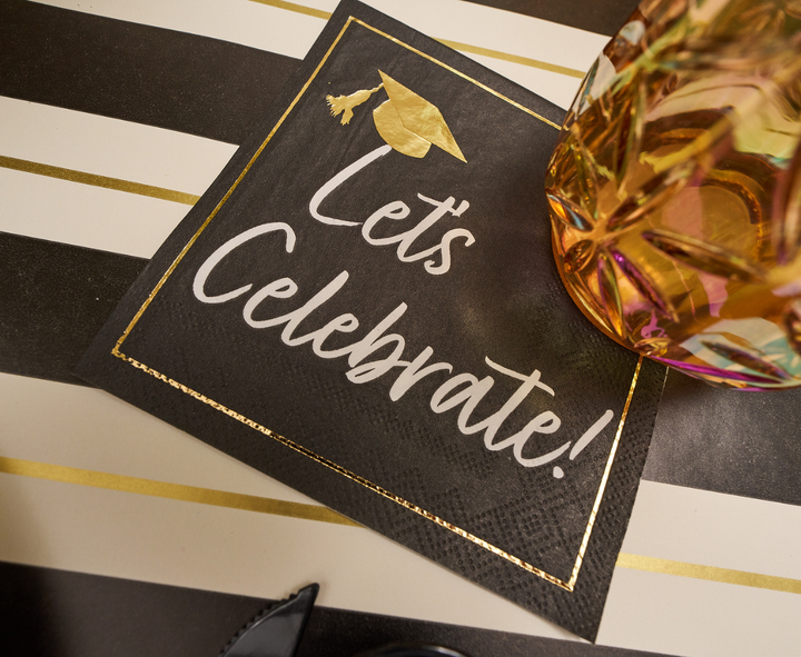 Sophistiplate’s Classic Grad Cocktail Napkins/26pk feature a black design with gold trim and “Let’s Celebrate!” cap motif. Shown on stripes with an amber-pink glass, these napkins are ideal for adding style to your graduation party.