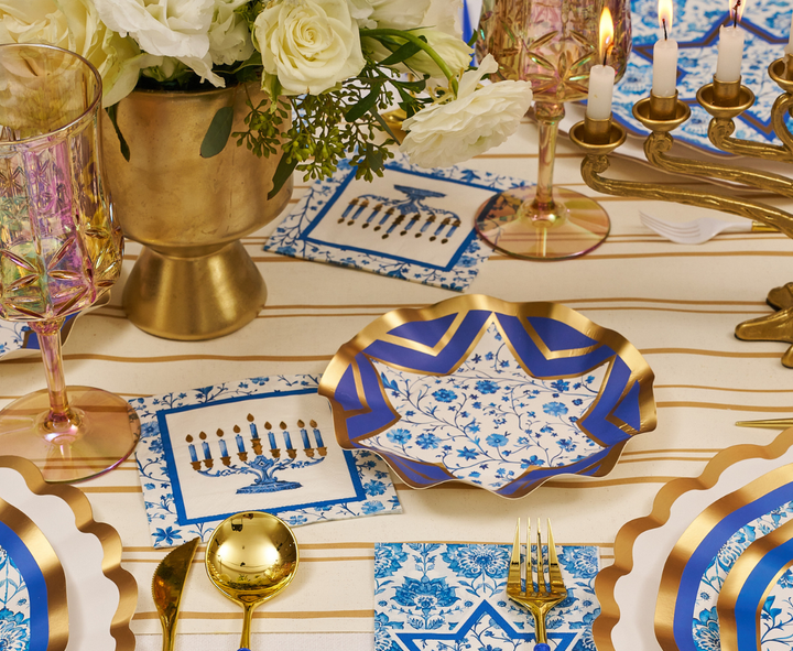 The Hanukkah Toile Table Setting by Sophistiplate features blue and gold designer paper plates, gold flatware, pink glasses, white flowers displayed in a gold vase, menorah napkins, and glowing candles atop a striped tablecloth—creating a festive tablescape that’s perfect for Hanukkah celebrations.