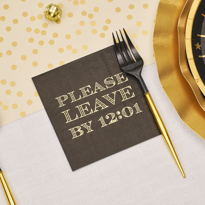 A black Midnight Sparkle Paper Cocktail Napkin Please Leave By 12:01 /26pk printed with the words PLEASE LEAVE BY 12:01 in gold, next to a gold-handled fork and layered plates.