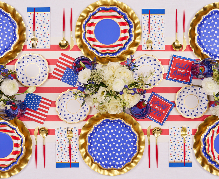 A festive table set with Sophistiplate's Party in the USA Guest Towels, themed plates and cups, gold cutlery, and red, white, and blue decor. White flowers and mini American flags complete this patriotic party look.