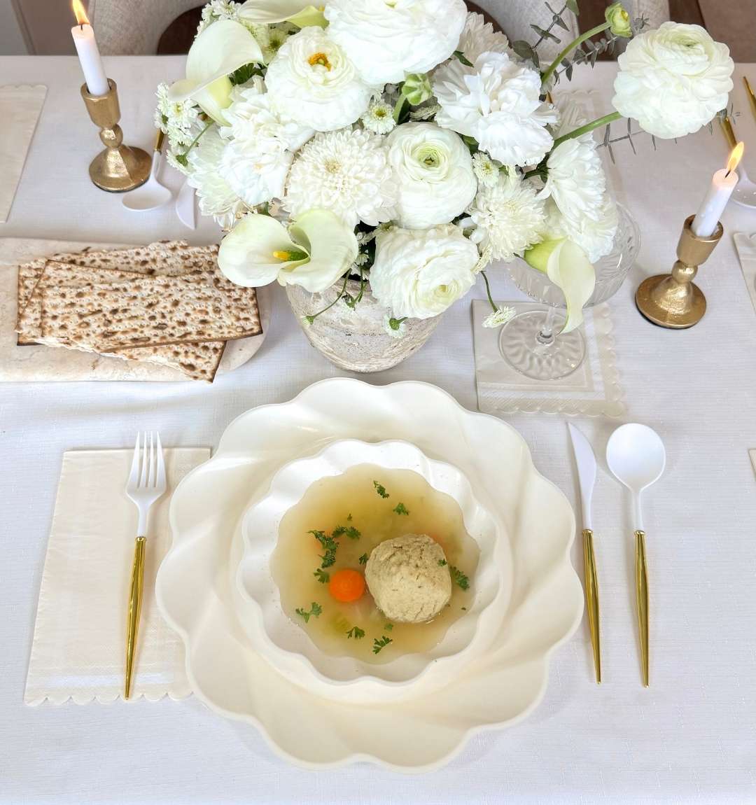 A white table featuring the Sophistiplate Eco-Friendly Table Setting- Cream: gold cutlery, stacked bamboo plates, matzo ball soup with carrots and herbs, matzo crackers, candles, and a white floral centerpiece.