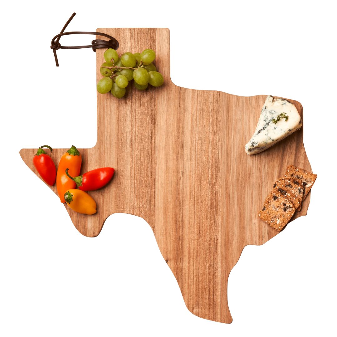 A wooden serving board shaped like Texas holds green grapes, colorful mini peppers, a wedge of blue cheese, and crackers.