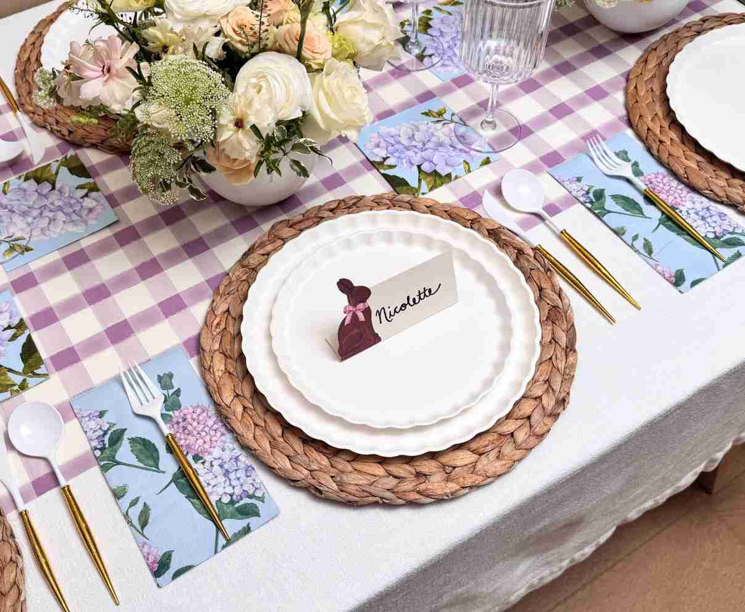 The Ultimate Easter Tablescape for 16: Violet Hydrangea by Sophistiplate LLC features woven placemats, designer disposable plates, floral napkins, gold cutlery, a “Nicolette” place card, and a white-pink floral centerpiece on a lavender-checkered runner.